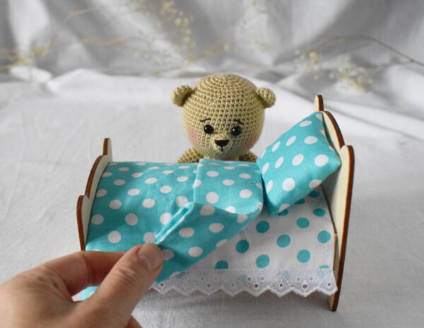 Soft toy bear beige color. Next to a wooden bed with a set of bed linen. On the bed there is a mattress, a sheet, a pillow, a blanket.