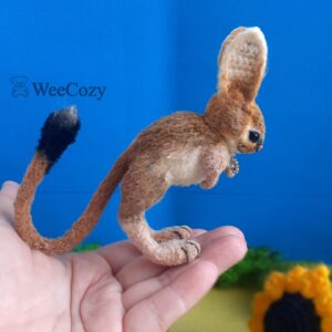 Crochet miniature realistic rodent jerboa stands on half-bent hind legs, the head is tilted down, the tail with a black tassel is bent up.