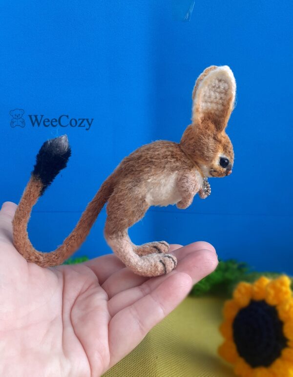 Crochet miniature realistic rodent jerboa stands on half-bent hind legs, the head is tilted down, the tail with a black tassel is bent up.