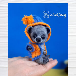Gray miniature crochet bear toy sits on the palm. He wears a blue and orange coat with a hood over his head.