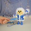 Soft toy winter hedgehog-girl. It stands on its own. White hairstyle. Two pigtails. Blue coat and boots. There is a small sled next to it. Knitted snowflakes for home decor are lying on the sled.