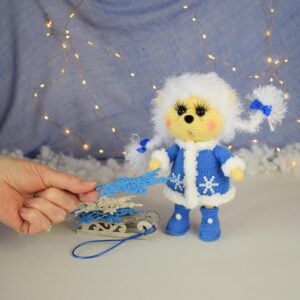 Soft toy winter hedgehog-girl. It stands on its own. White hairstyle. Two pigtails. Blue coat and boots. There is a small sled next to it. Knitted snowflakes for home decor are lying on the sled.