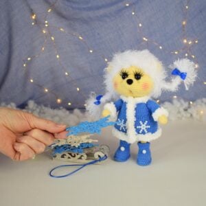 Hérisson d'hiver en peluche. Elle se tient debout toute seule. Coiffure blanche. Deux nattes. Manteau et bottes bleus. Il y a un petit traîneau à côté d'elle. Des flocons de neige tricotés pour la décoration intérieure sont posés sur le traîneau.