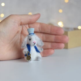 Miniature white rabbit. A soft toy. On his head is a light blue hat. A blue vest. He holds a watch pendant in his paw.