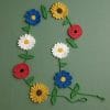 A handmade crochet wildflower garland featuring sunflowers, daisies, poppies, and blue blossoms with green leafy vines. The floral decoration is laid flat on a green background, perfect for spring or botanical-themed home decor.