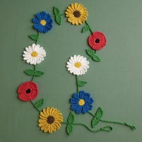 A handmade crochet wildflower garland featuring sunflowers, daisies, poppies, and blue blossoms with green leafy vines. The floral decoration is laid flat on a green background, perfect for spring or botanical-themed home decor.