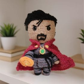 A handmade crochet doll inspired by Doctor Strange from Marvel. The doll features a dark blue outfit, red flowing cape, white side-streaked hair, and a magical medallion on the chest. A plush amigurumi version of the Sorcerer Supreme posed standing upright.