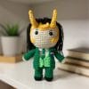 A handmade crochet doll based on President Loki from the Marvel Loki series. The doll features a green suit with a striped tie, black hair with tiny horns on a headband, and a mischievous expression. Inspired by the multiverse version of Loki, this plush amigurumi captures his chaotic charm.