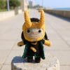 A handmade crochet doll inspired by Loki from Marvel comics and movies. The character features black hair, green and gold outfit details, a small horned helmet, and expressive embroidered eyes. The doll is posed upright on a shelf with neutral decor, highlighting its intricate craftsmanship and soft texture.