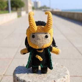 A handmade crochet doll inspired by Loki from Marvel comics and movies. The character features black hair, green and gold outfit details, a small horned helmet, and expressive embroidered eyes. The doll is posed upright on a shelf with neutral decor, highlighting its intricate craftsmanship and soft texture.