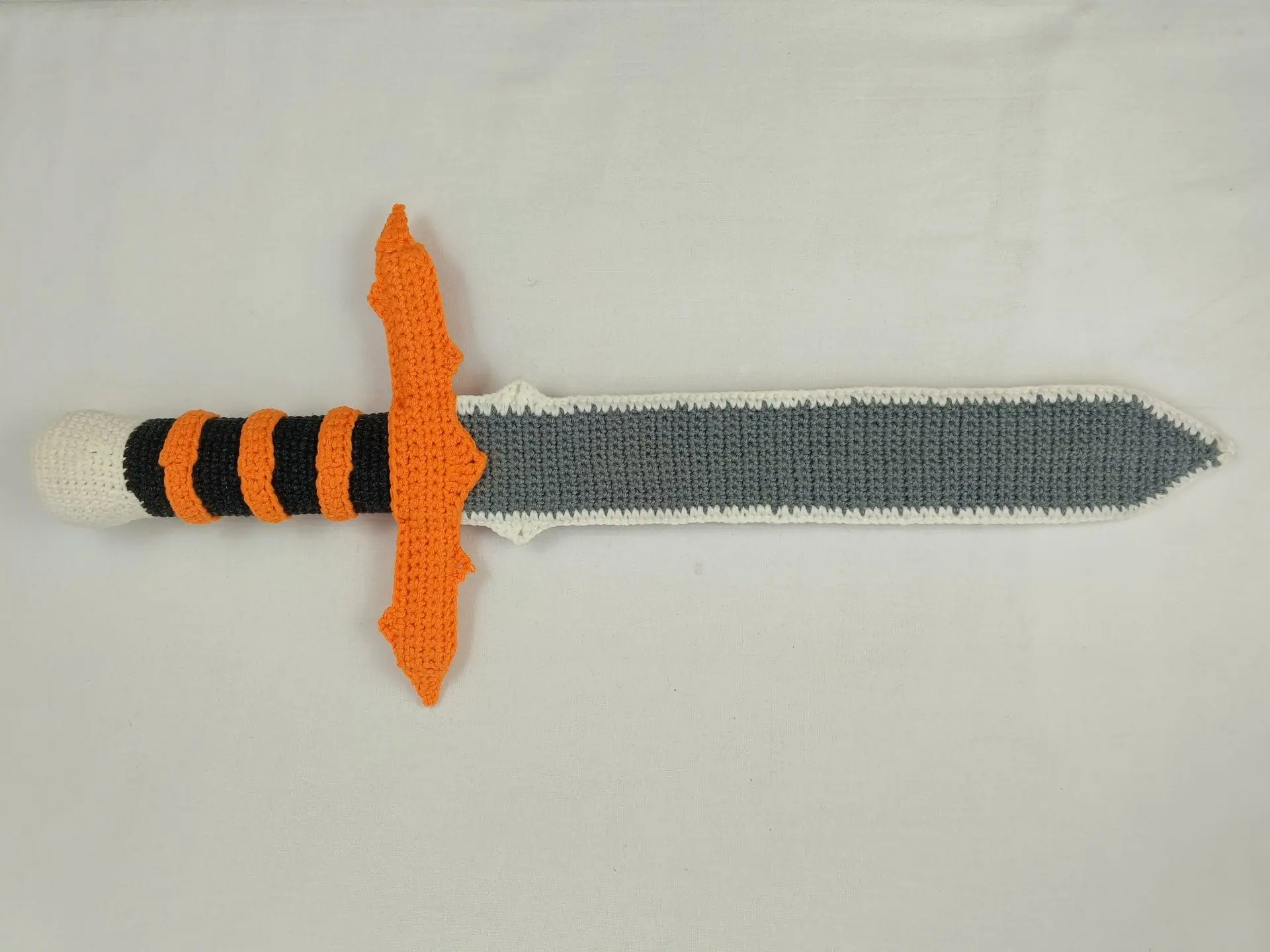 Halloween Crochet Sword Pattern – Skull and Spider Amigurumi Toy 9 Crocheted Halloween toy sword and sheath made with amigurumi technique. The sword has a skull handle, black-and-orange striped grip, and bat wing-shaped guard. The matching orange sheath features a black spider design. A soft, safe toy for kids, ideal for Halloween costumes, cosplay, or spooky-themed gifts. Clear handmade stitches and playful design.