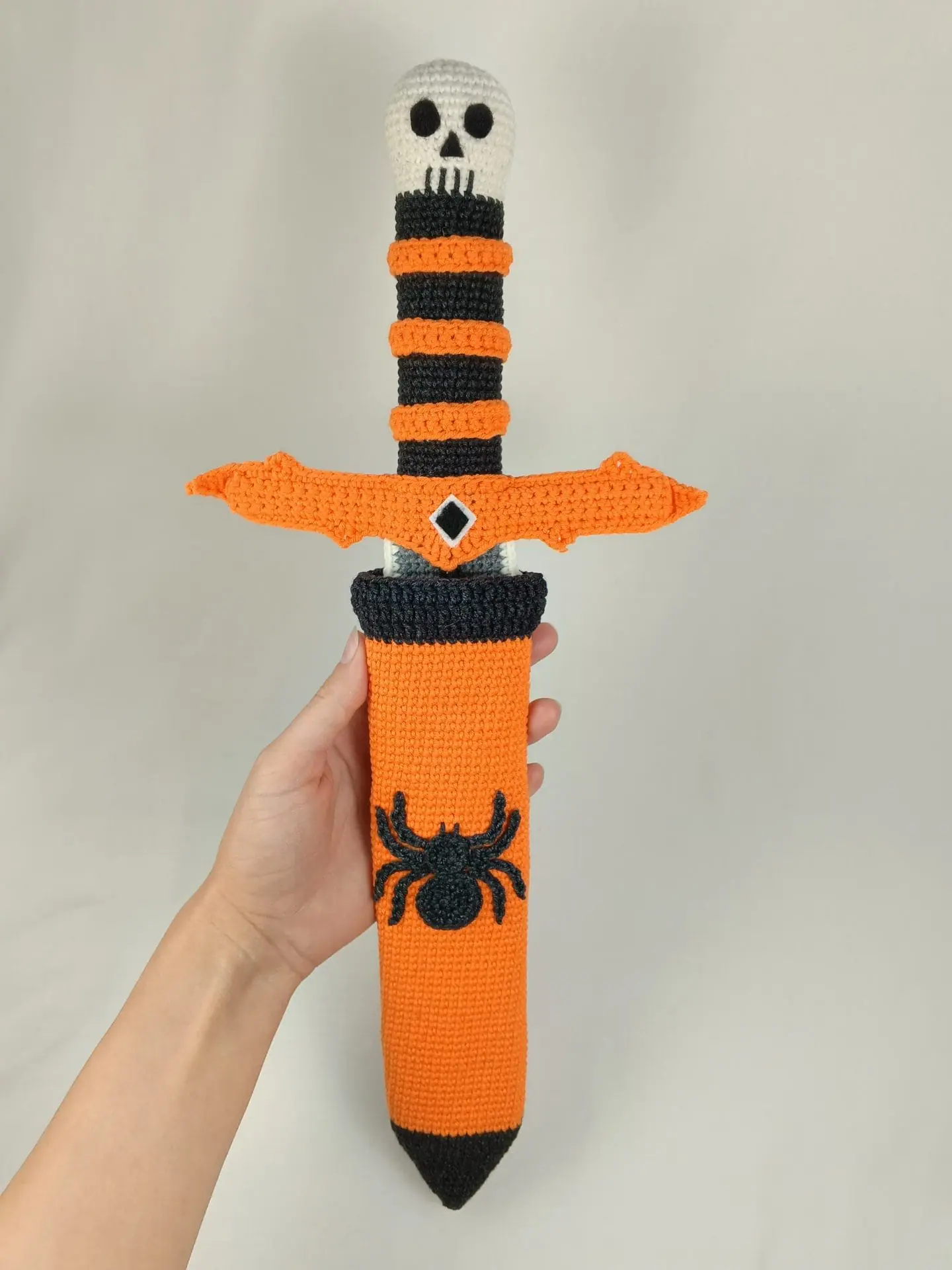 Halloween Crochet Sword Pattern – Skull and Spider Amigurumi Toy 1 Crocheted Halloween toy sword and sheath made with amigurumi technique. The sword has a skull handle, black-and-orange striped grip, and bat wing-shaped guard. The matching orange sheath features a black spider design. A soft, safe toy for kids, ideal for Halloween costumes, cosplay, or spooky-themed gifts. Clear handmade stitches and playful design.