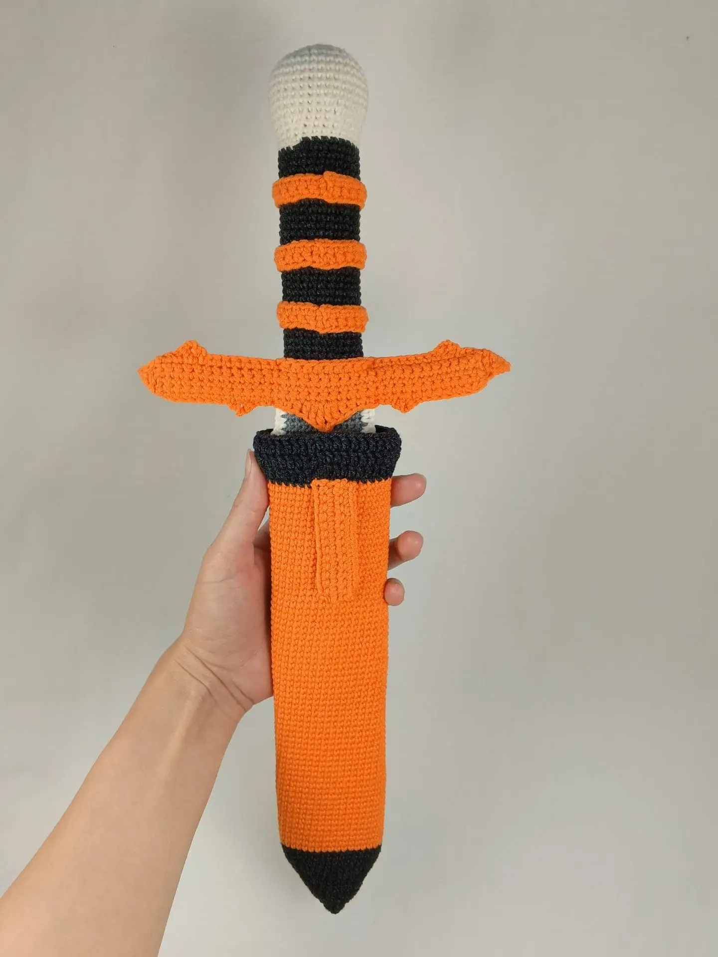 Halloween Crochet Sword Pattern – Skull and Spider Amigurumi Toy 2 Crocheted Halloween toy sword and sheath made with amigurumi technique. The sword has a skull handle, black-and-orange striped grip, and bat wing-shaped guard. The matching orange sheath features a black spider design. A soft, safe toy for kids, ideal for Halloween costumes, cosplay, or spooky-themed gifts. Clear handmade stitches and playful design.
