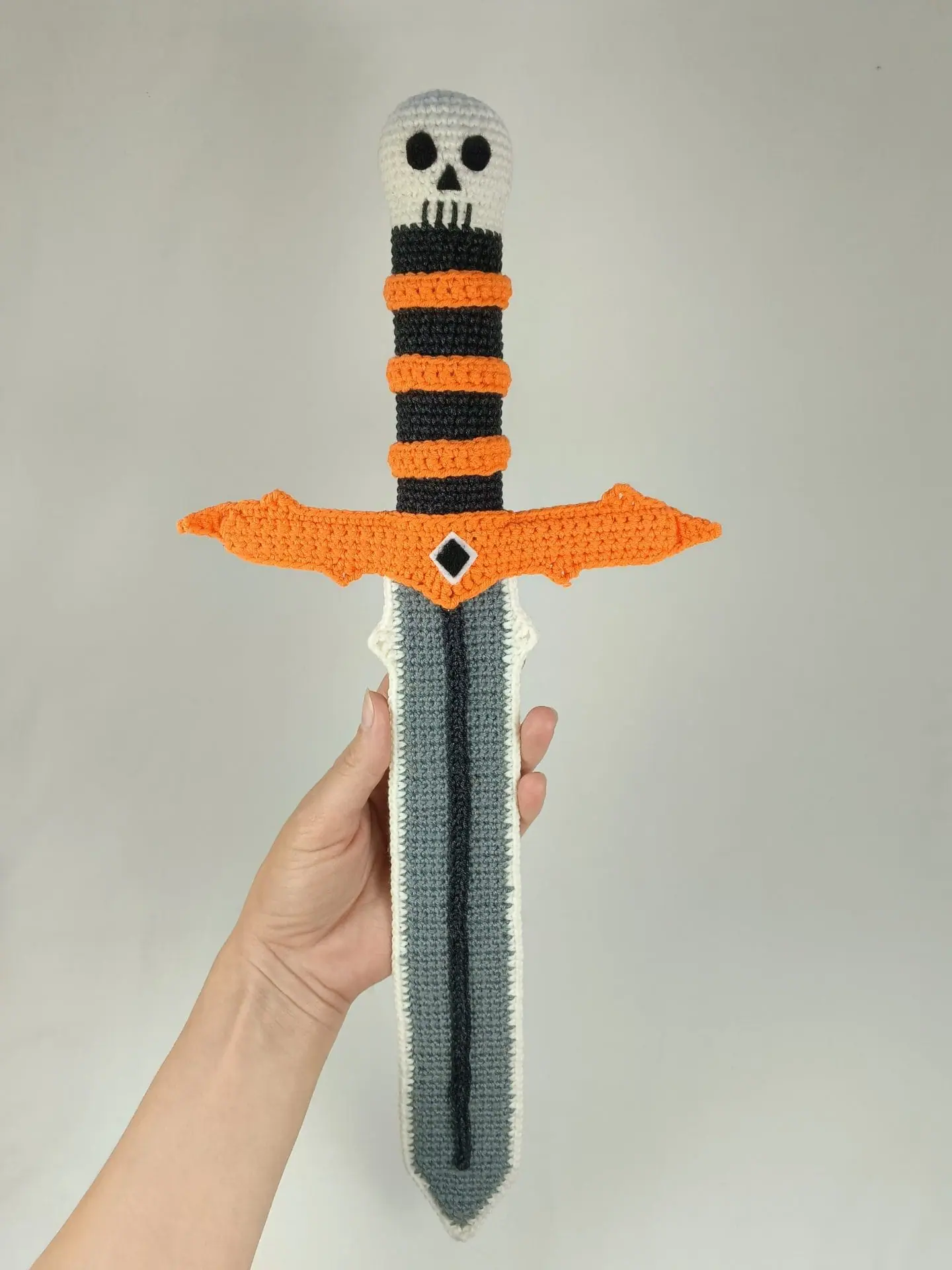 Halloween Crochet Sword Pattern – Skull and Spider Amigurumi Toy 3 Crocheted Halloween toy sword and sheath made with amigurumi technique. The sword has a skull handle, black-and-orange striped grip, and bat wing-shaped guard. The matching orange sheath features a black spider design. A soft, safe toy for kids, ideal for Halloween costumes, cosplay, or spooky-themed gifts. Clear handmade stitches and playful design.