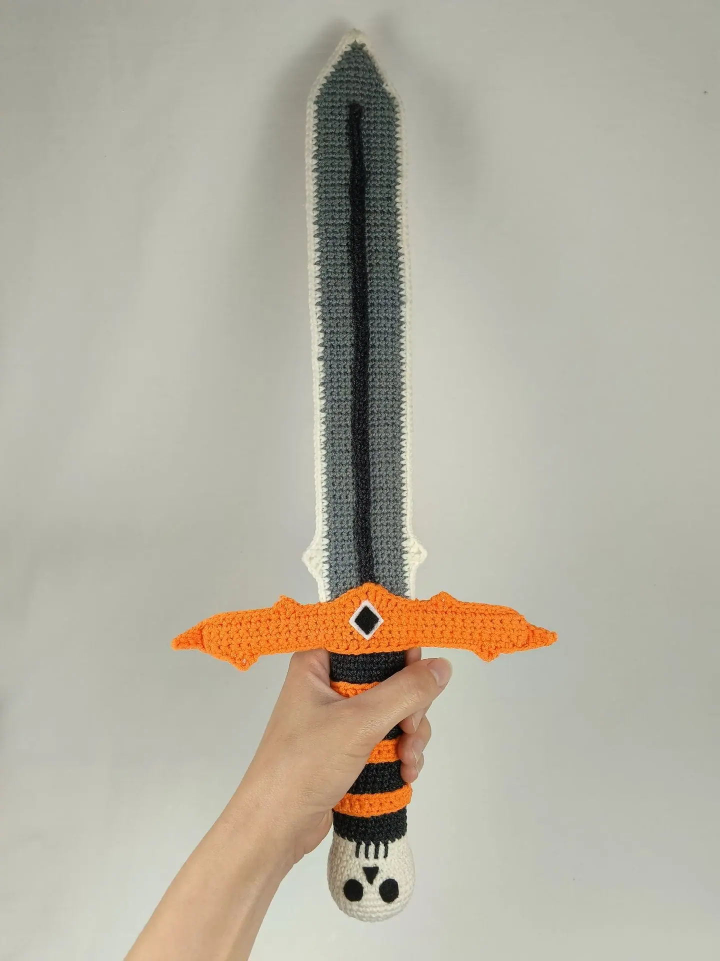 Halloween Crochet Sword Pattern – Skull and Spider Amigurumi Toy 4 Crocheted Halloween toy sword and sheath made with amigurumi technique. The sword has a skull handle, black-and-orange striped grip, and bat wing-shaped guard. The matching orange sheath features a black spider design. A soft, safe toy for kids, ideal for Halloween costumes, cosplay, or spooky-themed gifts. Clear handmade stitches and playful design.