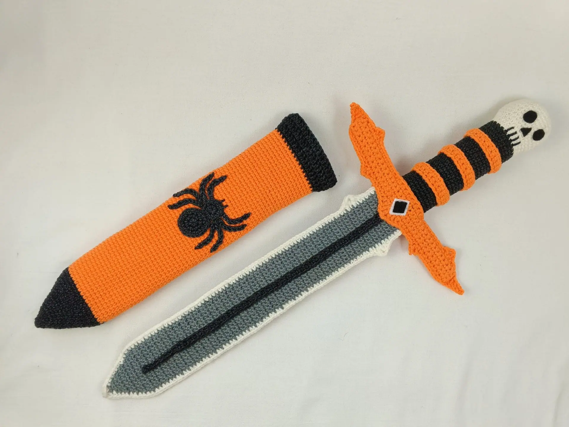 Halloween Crochet Sword Pattern – Skull and Spider Amigurumi Toy 6 Crocheted Halloween toy sword and sheath made with amigurumi technique. The sword has a skull handle, black-and-orange striped grip, and bat wing-shaped guard. The matching orange sheath features a black spider design. A soft, safe toy for kids, ideal for Halloween costumes, cosplay, or spooky-themed gifts. Clear handmade stitches and playful design.