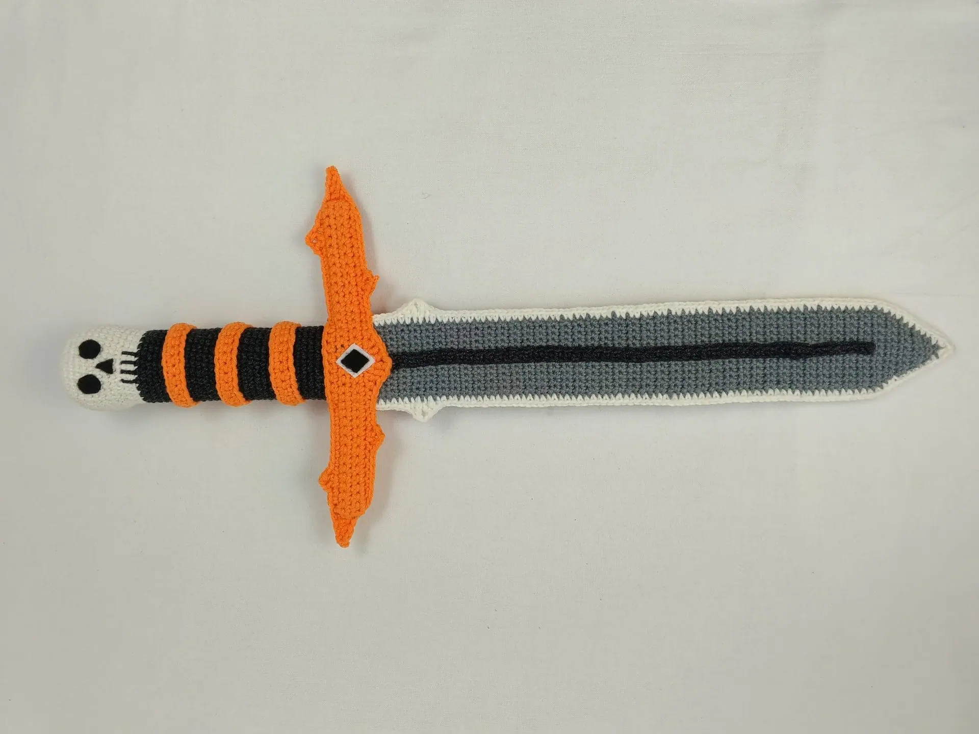Halloween Crochet Sword Pattern – Skull and Spider Amigurumi Toy 8 Crocheted Halloween toy sword and sheath made with amigurumi technique. The sword has a skull handle, black-and-orange striped grip, and bat wing-shaped guard. The matching orange sheath features a black spider design. A soft, safe toy for kids, ideal for Halloween costumes, cosplay, or spooky-themed gifts. Clear handmade stitches and playful design.