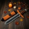 Crocheted Halloween toy sword and sheath made with amigurumi technique. The sword has a skull handle, black-and-orange striped grip, and bat wing-shaped guard. The matching orange sheath features a black spider design. A soft, safe toy for kids, ideal for Halloween costumes, cosplay, or spooky-themed gifts. Clear handmade stitches and playful design.