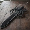 Crocheted sword with a black spider and white web design, handmade amigurumi toy on a neutral background.